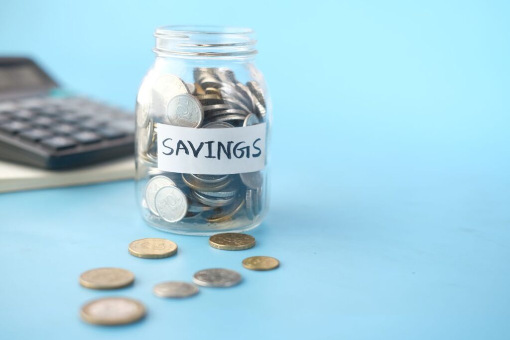 Glass jar labeled Savings filled with coins beside a calculator, representing Roth versus Traditional IRA tax planning